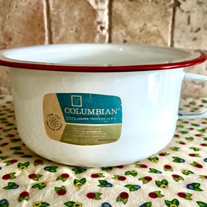 May include: White enamelware bowl with a red rim and a handle. The bowl has a label that reads "COLUMBIAN TERRE HAUTE INDIANA U.S.A." The bowl is sitting on a floral patterned fabric.