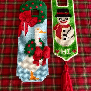 Op de afbeelding: Twee kerstdeurhangers. Eén toont een gans met een krans en strik, de andere een sneeuwpop met een hoed en sjaal. Beide zijn gemaakt van kruissteek met rode, groene, witte en blauwe kleuren. Eén heeft een rode kwast.