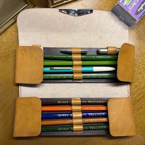 Op de afbeelding: Een beige lederen etui met twee binnenvakken en elastische lussen. Het bovenste gedeelte bevat vier groene en turquoise schrijfinstrumenten. Het onderste gedeelte bevat vijf potloden met de tekst Seiner Kraft Triumph erop gedrukt.
