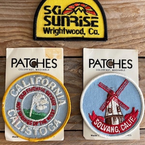 May include: Three embroidered patches on backing cards. One is a yellow semi-circle with a sunrise graphic and text reading SG SUNRISE Wrightwood, Ca. Two are circular: one light blue with a geyser and text reading CALIFORNIA CALISTOGA, and one light blue with a windmill and text reading SOLVANG CALIF.