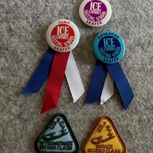 May include: Four Ice Capades Skating School badges with ribbons and two triangular patches. The badges are red, blue, purple, and light blue with white lettering. The patches are green and yellow with white lettering. The badges say "Jump", "Spin", "Advanced", and "Ice Capades Skating School Skater". The patches say "Intermediate Second Class" and "Novice Second Class".