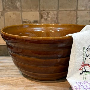 May include: A large, brown ceramic mixing bowl with horizontal ridges. A white cloth with embroidered details is partially visible on the right side. The bowl sits on a wooden surface.
