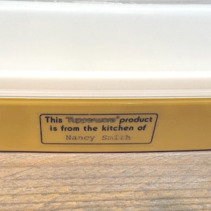May include: A close-up of a Tupperware container with a yellow base and a white lid. The text on the container reads: "This Tupperware product is from the kitchen of Nancy Smith."