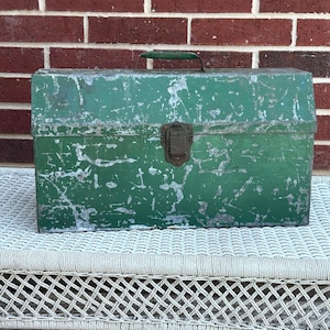 May include: A vintage green metal toolbox with a handle and a latch. The toolbox has a distressed, weathered appearance with visible wear and tear. It sits on a white wicker surface against a red brick wall.