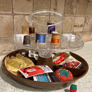 Peut inclure: Un organisateur de couture clair à deux niveaux avec des bobines de fil de différentes couleurs, un dé à coudre et des coussins à épingles. Le niveau inférieur contient des coussins à épingles et des paquets de fournitures de couture. L'organisateur est marron et transparent.