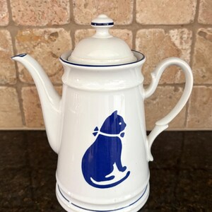 May include: White ceramic teapot with a blue cat silhouette design. The teapot has a curved spout, a handle, and a lid with a small knob. Blue trim accents the edges. A charming addition to any kitchen.