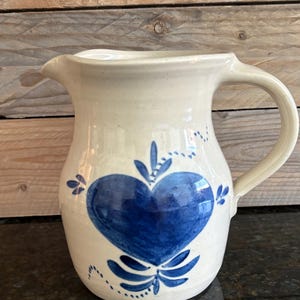 May include: Cream-colored ceramic pitcher with a large blue heart design. The heart is centered on the front and surrounded by blue floral accents. The pitcher has a curved handle and a spout for pouring.