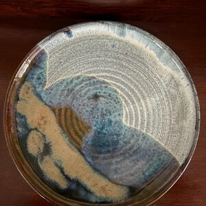 May include: A handmade ceramic plate with a unique, organic design. The plate features a blend of blue, tan, and gray hues, with a glossy finish. The circular shape and textured surface add to its artistic appeal. This decorative plate is suitable for serving food or as a display piece.
