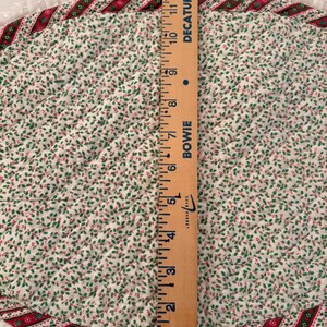 May include: An oval quilted placemat featuring a white fabric with a small green and red holly leaf pattern. The edge is trimmed with a red and green striped border and a white ruffled fringe. A wooden ruler shows a length of 13 inches.