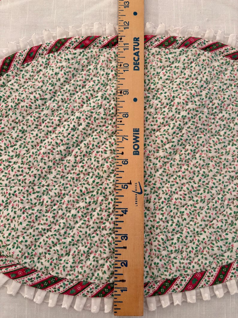 May include: An oval quilted placemat featuring a white fabric with a small green and red holly leaf pattern. The edge is trimmed with a red and green striped border and a white ruffled fringe. A wooden ruler shows a length of 13 inches.