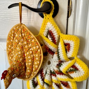 May include: A pair of decorative kitchen items: a quilted chicken-shaped oven mitt and a yellow, white, and red crocheted pot holder. The mitt features a yellow floral pattern and a red comb. The pot holder has a sunflower design.