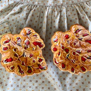 May include: Two leaf-shaped ceramic dishes with a glossy finish. The dishes have a base color of tan with red and brown accents. The dishes are on a patterned fabric background.