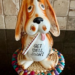 May include: A ceramic dog figurine with a thermometer in its mouth and the words "GET WELL SOON!" on its chest. The dog has brown and white fur, long ears, and is sitting on a woven mat with a colorful braided edge.