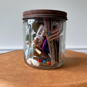 May include: A clear glass jar filled with various small objects, including a miniature American flag, a small bottle, and other trinkets. The jar has a rusty metal lid and sits on a brown surface. The contents are visible through the glass.