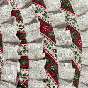 May include: A set of circular fabric placemats featuring a festive pattern of red and green holly leaves and berries on a white background. Each placemat is finished with a gathered white ruffled trim along the outer edge, adding texture.
