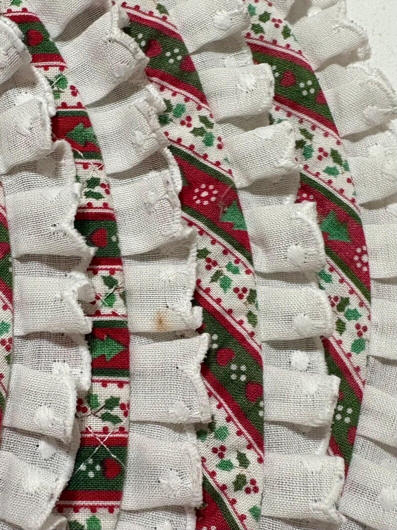 May include: A set of circular fabric placemats featuring a festive pattern of red and green holly leaves and berries on a white background. Each placemat is finished with a gathered white ruffled trim along the outer edge, adding texture.