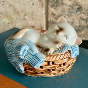 May include: A ceramic figurine of a sleeping cat nestled in a woven basket. The cat is white with brown markings and a pink nose, resting on a blue blanket. The basket is brown with small holes.