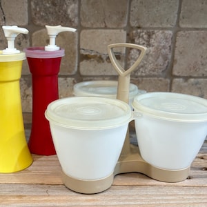 Könnte beinhalten: Eine Sammlung von Vintage-Küchenbehältern. Enthält einen gelben und roten Spender mit weißen Pumpköpfen und einen Doppelbehälter mit weißen Schalen und Deckeln sowie einen beigefarbenen Griff.