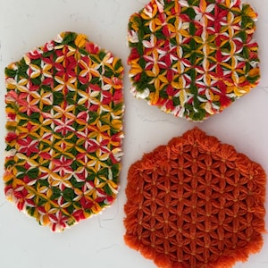 Op de afbeelding: Drie handgemaakte, zeshoekige onderzetters. Twee onderzetters hebben een patroon van rood, groen, geel en wit garen. De derde onderzetter is effen oranje. De onderzetters zijn gemaakt van garen en beschermen oppervlakken tegen hitte.