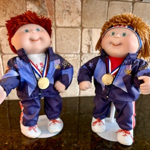 May include: Two Cabbage Patch Kids dolls dressed in matching blue tracksuits with red and white accents. Each doll wears a gold medal and has red hair. The dolls are standing on white bases.