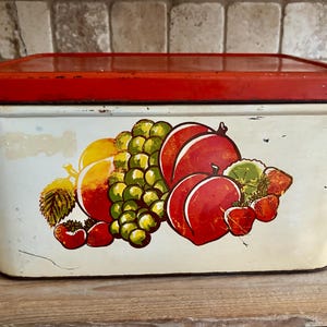 May include: Vintage rectangular metal bread box with a red lid and white body. The box features a colorful illustration of fruits, including peaches, grapes, and strawberries. The box is likely from the mid-20th century.