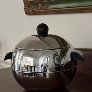 May include: A spherical chrome ice bucket featuring a polished silver finish with embossed penguin figures around the centre. The container includes two black side handles and a matching black spherical knob on the removable lid.