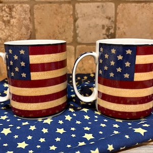 May include: Two ceramic mugs with a patriotic American flag design. The mugs feature a white handle and rim, with the flag's stars and stripes wrapping around the body. They sit on a blue cloth with yellow star patterns.