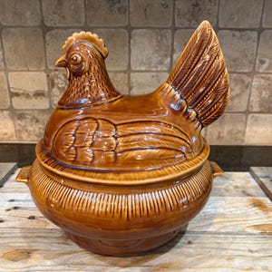 Könnte beinhalten: Eine braune Keramik-Hühner-Auflaufform mit Deckel. Die Schüssel hat ein detailliertes Design einer Henne mit Kamm, Schnabel und Flügeln. Der Boden hat vertikale Linien. Die Schüssel steht auf einer Holzoberfläche.