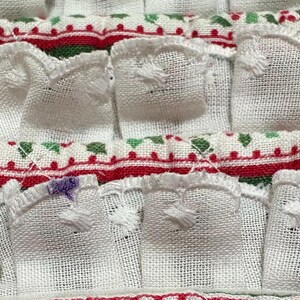 May include: A white fabric textile featuring rows of gathered ruffles with a woven texture. The horizontal bands are decorated with a repeating pattern of red and green geometric shapes and dots, with small purple embroidered accents on edges.