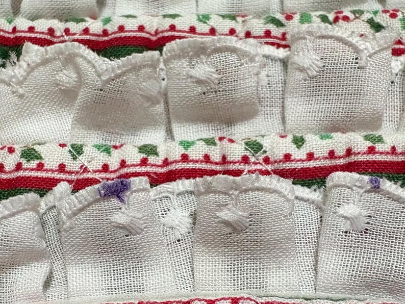 May include: A white fabric textile featuring rows of gathered ruffles with a woven texture. The horizontal bands are decorated with a repeating pattern of red and green geometric shapes and dots, with small purple embroidered accents on edges.