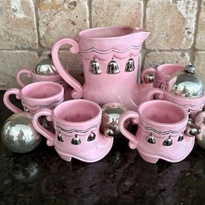 May include: A pink ceramic tea set featuring a boot-shaped pitcher and matching cups. The pitcher and cups are adorned with silver bell accents. Silver ornaments are placed around the set.