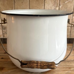 Könnte beinhalten: Weißer Emaille-Eimer mit schwarzem Rand und Metallgriff. Der Griff hat einen Holzgriff. Der Eimer steht auf einer Holzoberfläche. Dieser Vintage-Eimer ist ideal für die Einrichtung im Landhausstil.