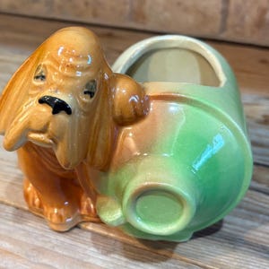 May include: A ceramic planter shaped like a brown basset hound dog with long ears and a black nose, attached to a cylindrical container with a glossy light green finish. The dog figure has a smooth, glazed surface and a hollow opening for plants.