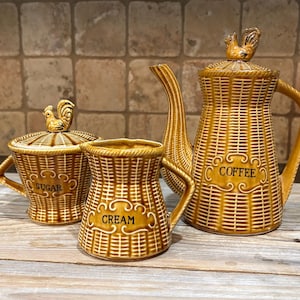 May include: A ceramic coffee service set in a golden-brown color with a textured basket-weave pattern. The set includes a coffee pot, a sugar bowl with a lid, and a creamer. Each piece features a decorative label with the words COFFEE, SUGAR, or CREAM.