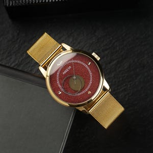 Puede incluir: Reloj de pulsera dorado con esfera roja y correa de malla. La esfera del reloj muestra la palabra "PAKETA" y un diseño circular. El reloj está sobre un fondo oscuro.