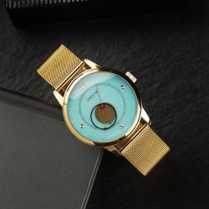 May include: Gold-toned wristwatch with a mesh band and a light blue face. The watch face features a circular design with a red accent and the word "PAKEТА". The watch is set against a dark background.