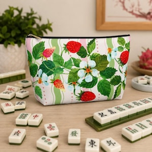 Könnte beinhalten: Eine weiße Kosmetiktasche mit Erdbeer- und Blumendesign. Die Tasche hat einen schwarzen Reißverschluss und ein Muster aus roten Erdbeeren, grünen Blättern und weißen Blüten. Die Tasche steht auf einer Holzoberfläche mit Mahjong-Steinen.