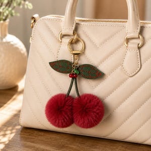 May include: A cherry-themed keychain with a gold-colored ring, green leaves, and two fluffy red pom-poms. The keychain is attached to a cream-colored handbag with a quilted pattern. The leaves have red stitching.
