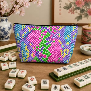 May include: A colorful cosmetic bag with a vibrant snakeskin pattern in pink, green, blue, purple, and yellow. The bag is black-zippered and sits among Mahjong tiles on a wooden surface. A vase of pink flowers is in the background.