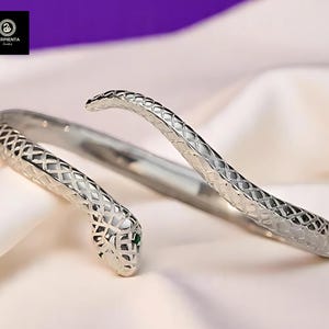 以下が含まれることがあります： 緑色の石の目をつけた銀色の蛇のブレスレット。ブレスレットは、手首に巻き付く蛇のようにデザインされています。