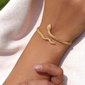 May include: Gold-coloured snake cuff bracelet. The bracelet wraps around the wrist with a textured, scale-like pattern. The snake's head and tail are visible, adding a unique design. The jewellery is worn on a person's arm.
