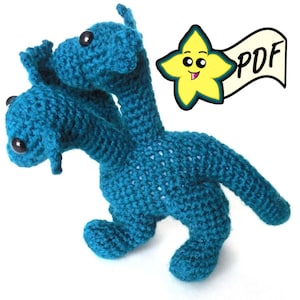 Può includere: Un giocattolo di peluche di drago a due teste di colore blu turchese lavorato a uncinetto. Il drago ha una coda lunga e due paia di gambe. Il giocattolo è realizzato con un punto a uncinetto testurizzato.