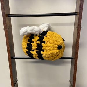May include: A crocheted bee-shaped plush toy with yellow and black stripes and white wings. The toy is hanging from a wooden shelf.
