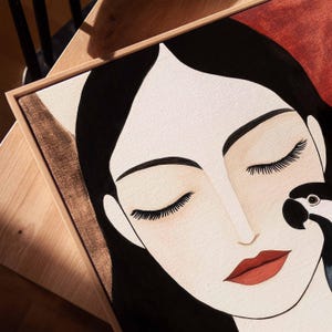 Modern abstract portrait of a woman with long black hair, closed eyes, and red lips, rendered in soft minimalist shapes. A bold blue parrot perches close to her cheek, its feathers textured in deep navy and soft sky blue. The neutral background of warm cream and muted rust tones adds warmth and balance, creating a tranquil scene of harmony between human and nature.