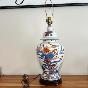 Könnte beinhalten: Eine Keramik-Tischlampe mit weißem Korpus und handgemaltem Blumenmuster in Blau-, Orange- und Goldtönen. Die Lampe hat die Form eines Ingwertopfes mit passendem Deckel, einer metallenen Lampenfassung und einem dunkelbraunen runden Fuß.