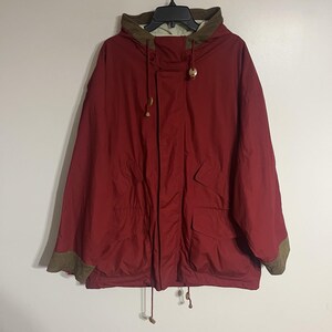 May include: A red hooded jacket with a brown collar and cuffs. The jacket has two front pockets and a drawstring at the waist. The hood has a drawstring with a wooden toggle.