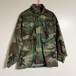 May include: Camouflage pattern military jacket with the text "U.S. ARMY" and a name tag. The jacket features multiple pockets and a button-up front. The color palette includes shades of green, brown, and black.