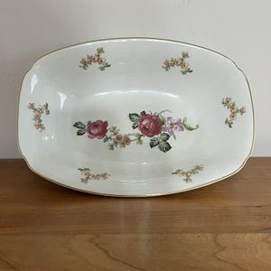 Puede incluir: Una fuente rectangular de cerámica blanquecina con borde dorado. La fuente está decorada con racimos de pequeñas flores amarillas y rosas, y un arreglo floral central de rosas rojas y flores moradas.