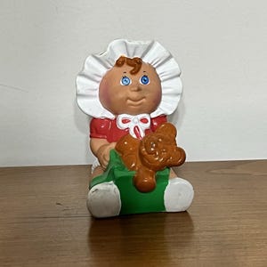 May include: A vintage Cabbage Patch Kids figurine of a baby wearing a red dress and white bonnet. The baby is holding a brown teddy bear and sitting on a green base. The figurine has blue eyes and a smiling expression.