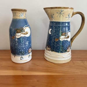 May include: A ceramic pitcher and vase set. The pitcher has a handle and a wide spout. Both pieces are painted with a winter scene, featuring a blue background with snowflakes, a snowman, and small houses. The rims are tan with blue snowflake designs.
